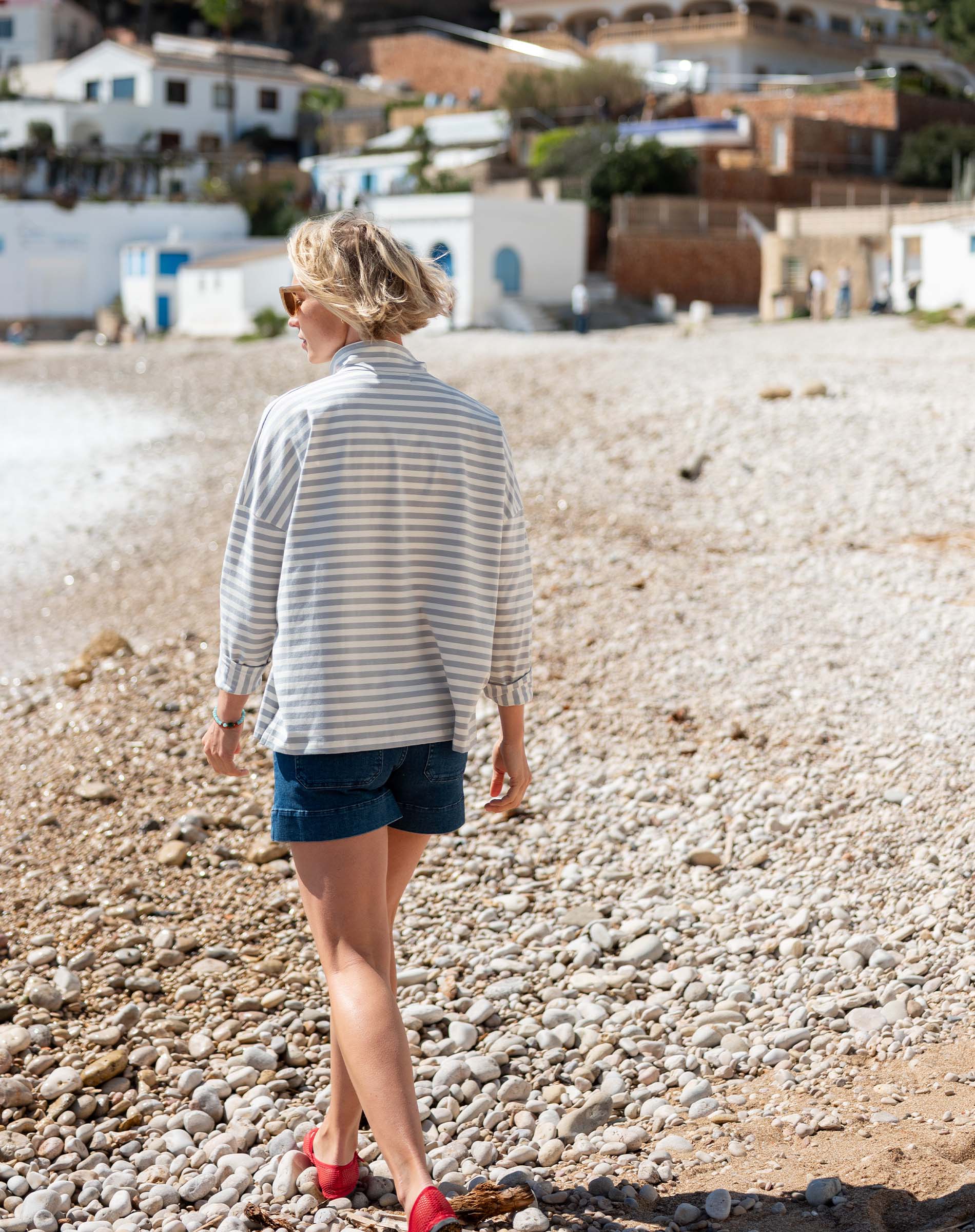 Woman in a striped shirt and shorts walking on a pebbly beach with buildings in the background.