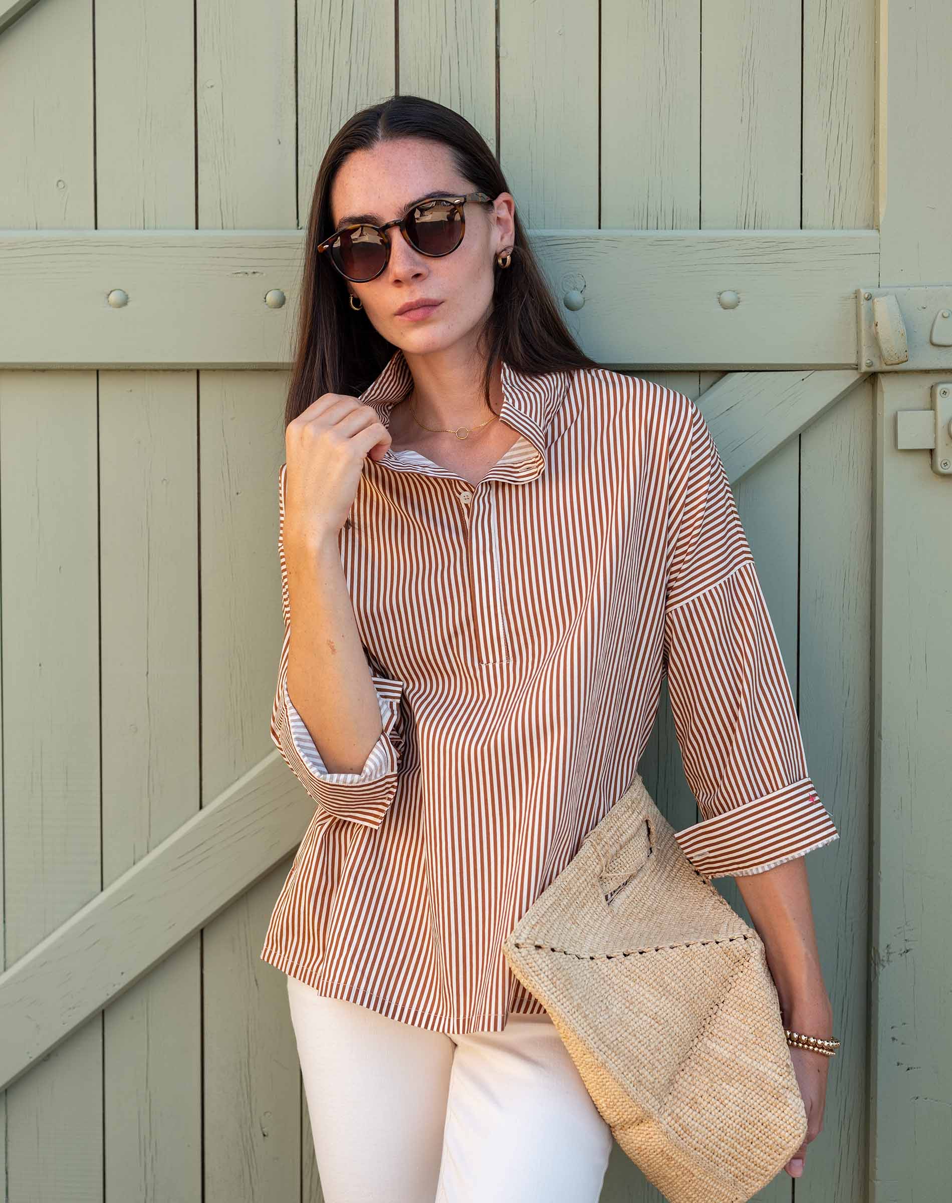 Woman in a striped shirt and sunglasses holding a beige bag against a wooden background