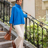 Woman in a blue jacket and white pants standing on outdoor steps with plants and a building in the background.