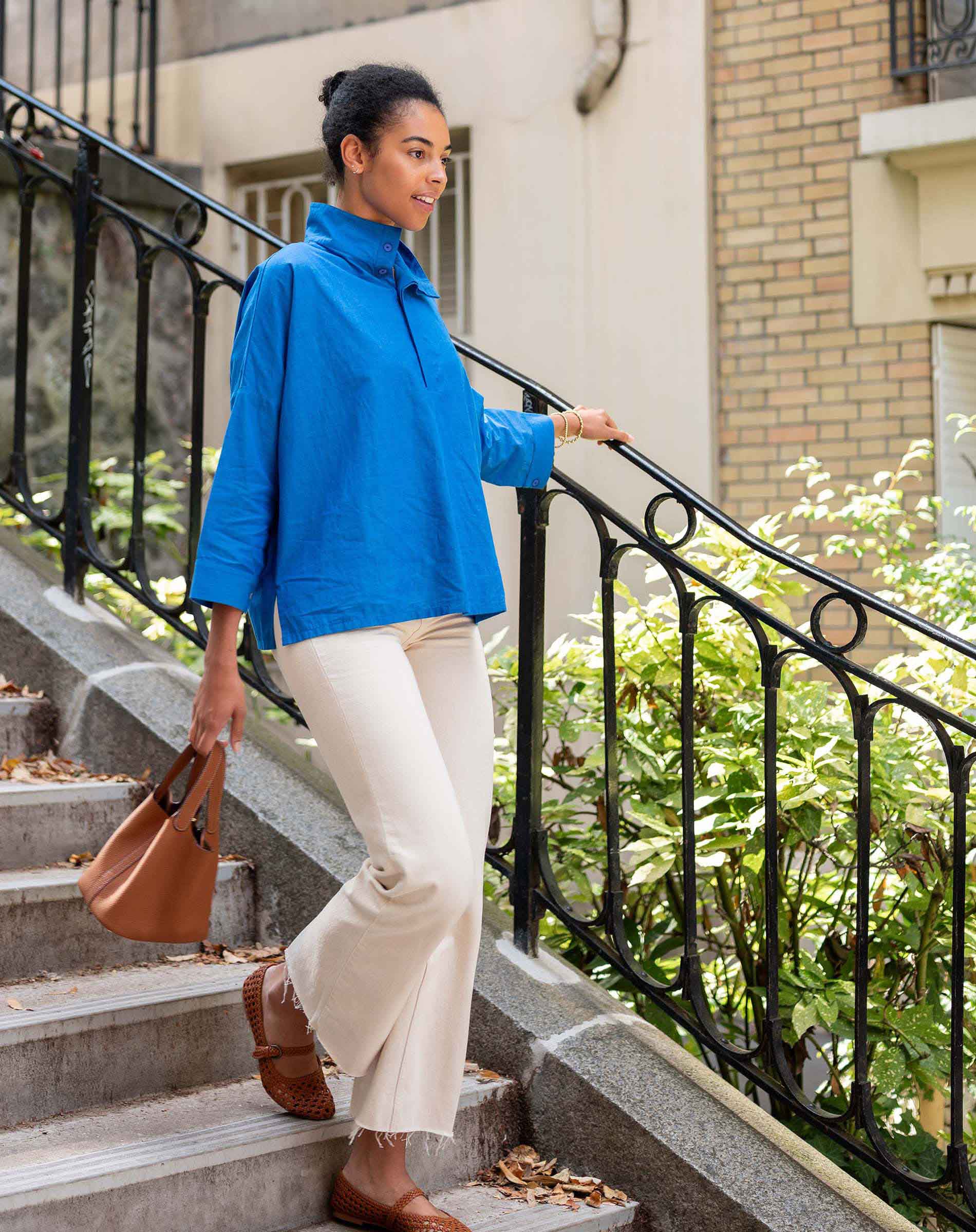 Woman in a blue jacket and white pants standing on outdoor steps with plants and a building in the background.