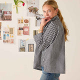 Woman wearing a black and white checkered shirt standing in front of a wall with various photos and notes.