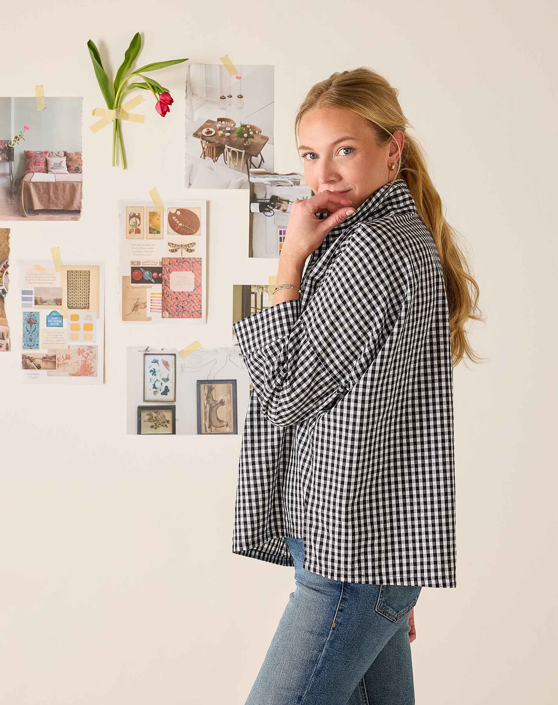Woman wearing a black and white checkered shirt standing in front of a wall with various photos and notes.