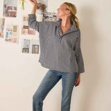 Woman wearing a checkered shirt and jeans, standing against a wall with decorative items.