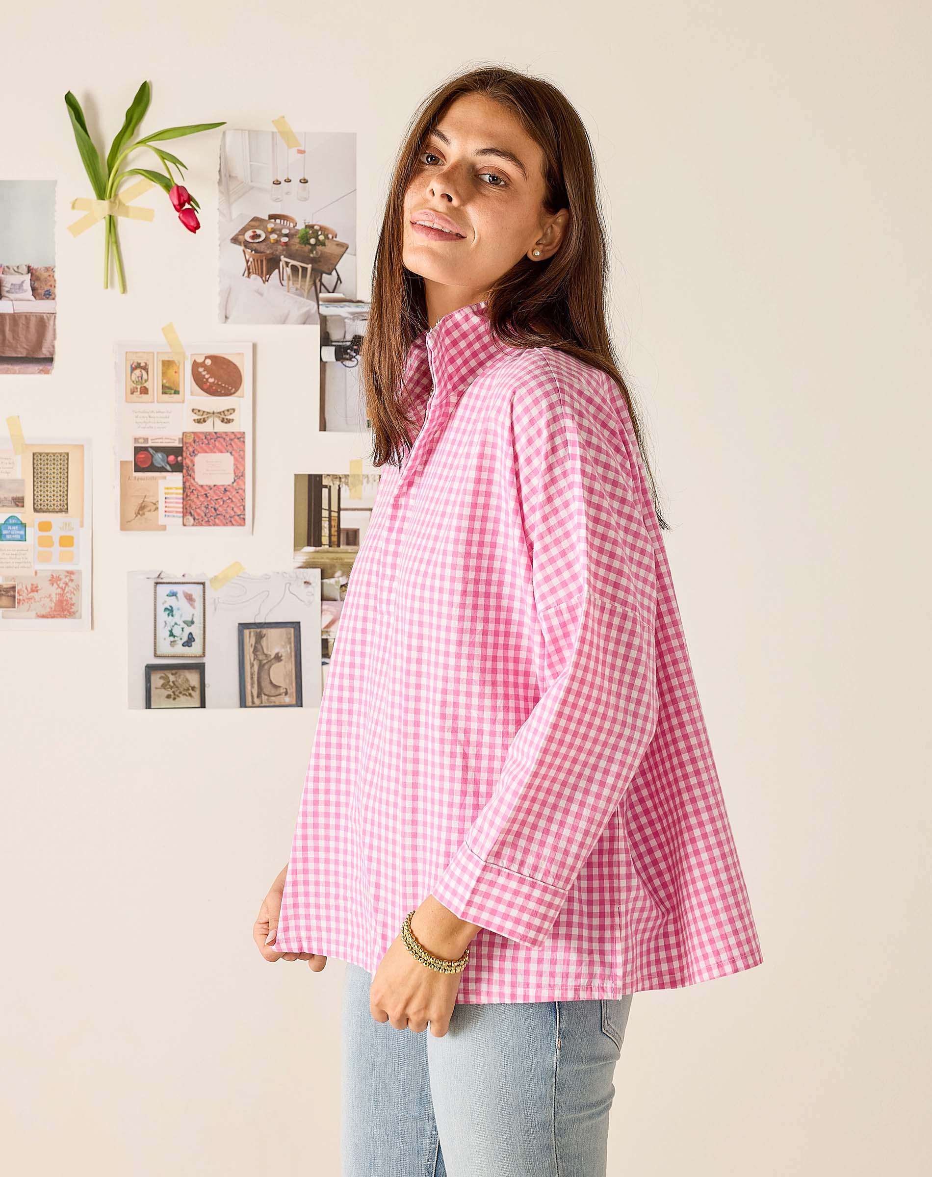 Woman wearing a pink checkered shirt standing in front of a wall with decorative items.