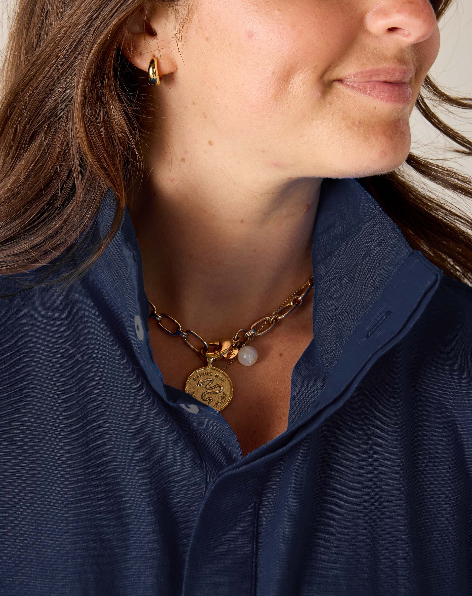 Close-up of a person wearing a gold necklace with a pendant and pearl, against a neutral background.