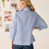 Woman wearing a blue striped shirt with rolled-up sleeves, standing in front of a wall with decorative items.