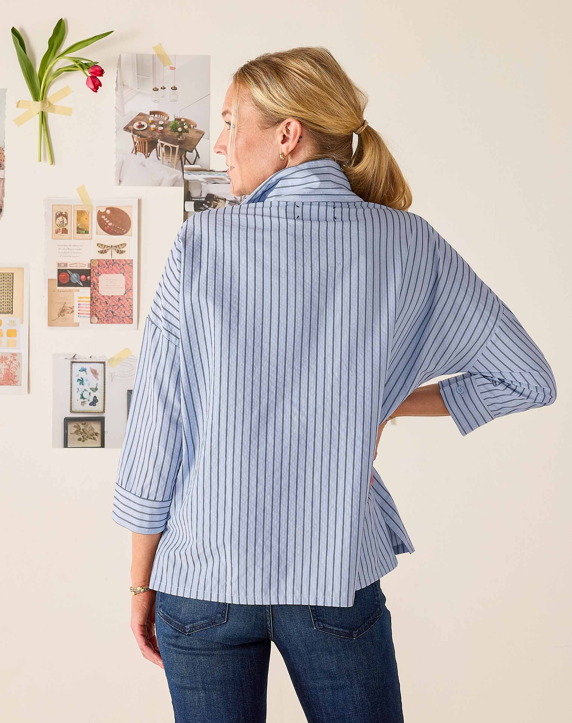 Woman wearing a blue striped shirt with rolled-up sleeves, standing in front of a wall with decorative items.