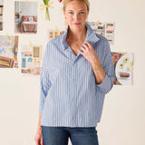 Woman wearing a blue striped shirt and jeans standing in front of a wall with various photos and decorations.