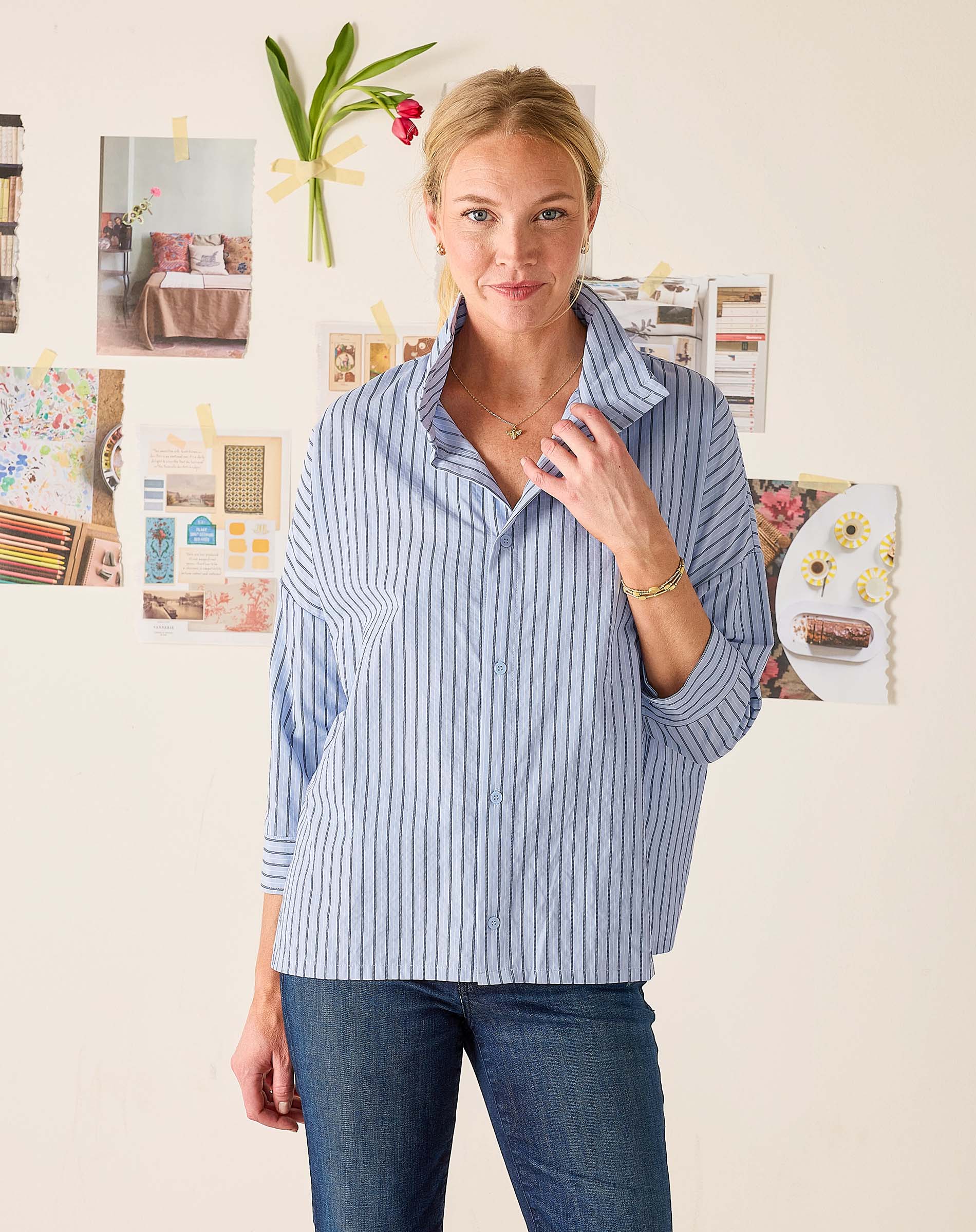 Woman wearing a blue striped shirt and jeans standing in front of a wall with various photos and decorations.