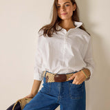 Woman wearing a white shirt and blue jeans with a woven belt against a plain background