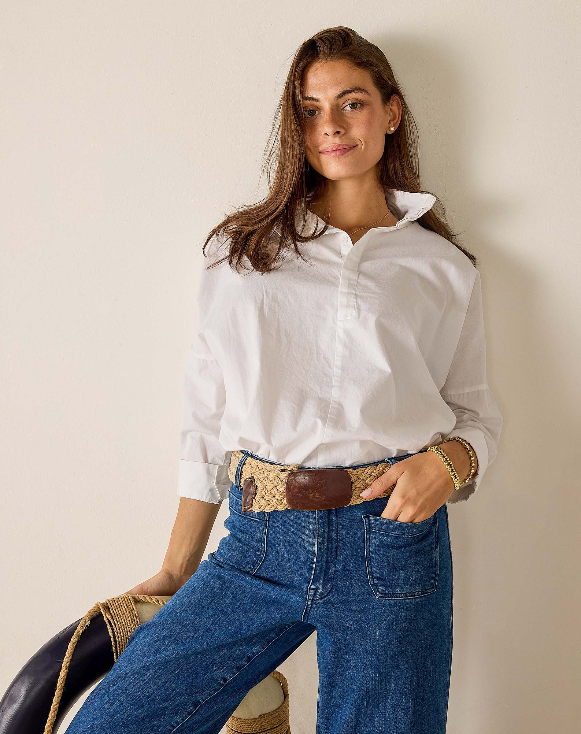 Woman wearing a white shirt and blue jeans with a woven belt against a plain background