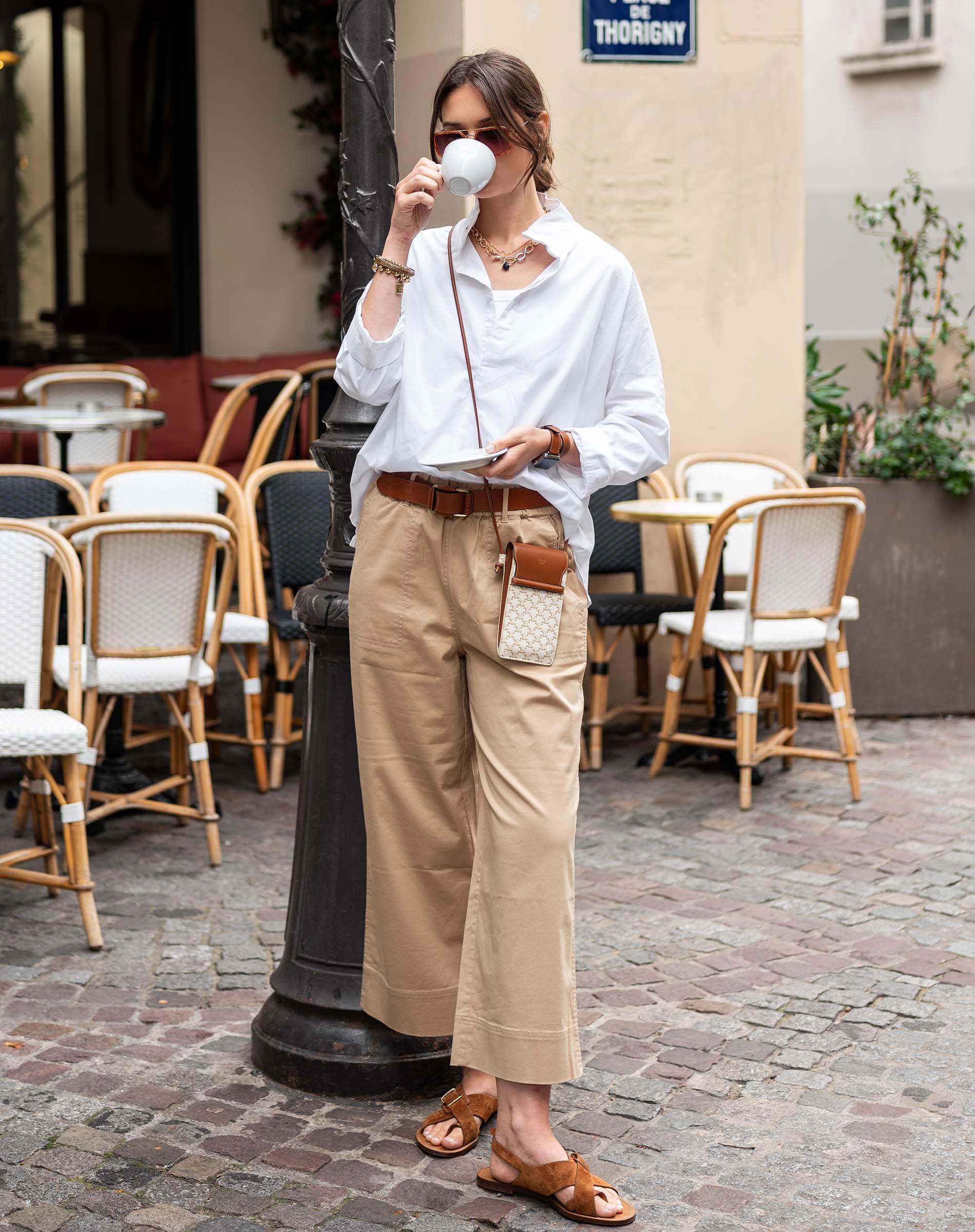 Woman in white shirt and beige pants standing on a street with cafe chairs in the background