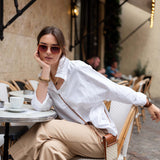 Woman sitting at an outdoor cafe wearing a white shirt and beige pants.
