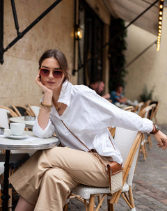 Woman sitting at an outdoor cafe wearing a white shirt and beige pants.