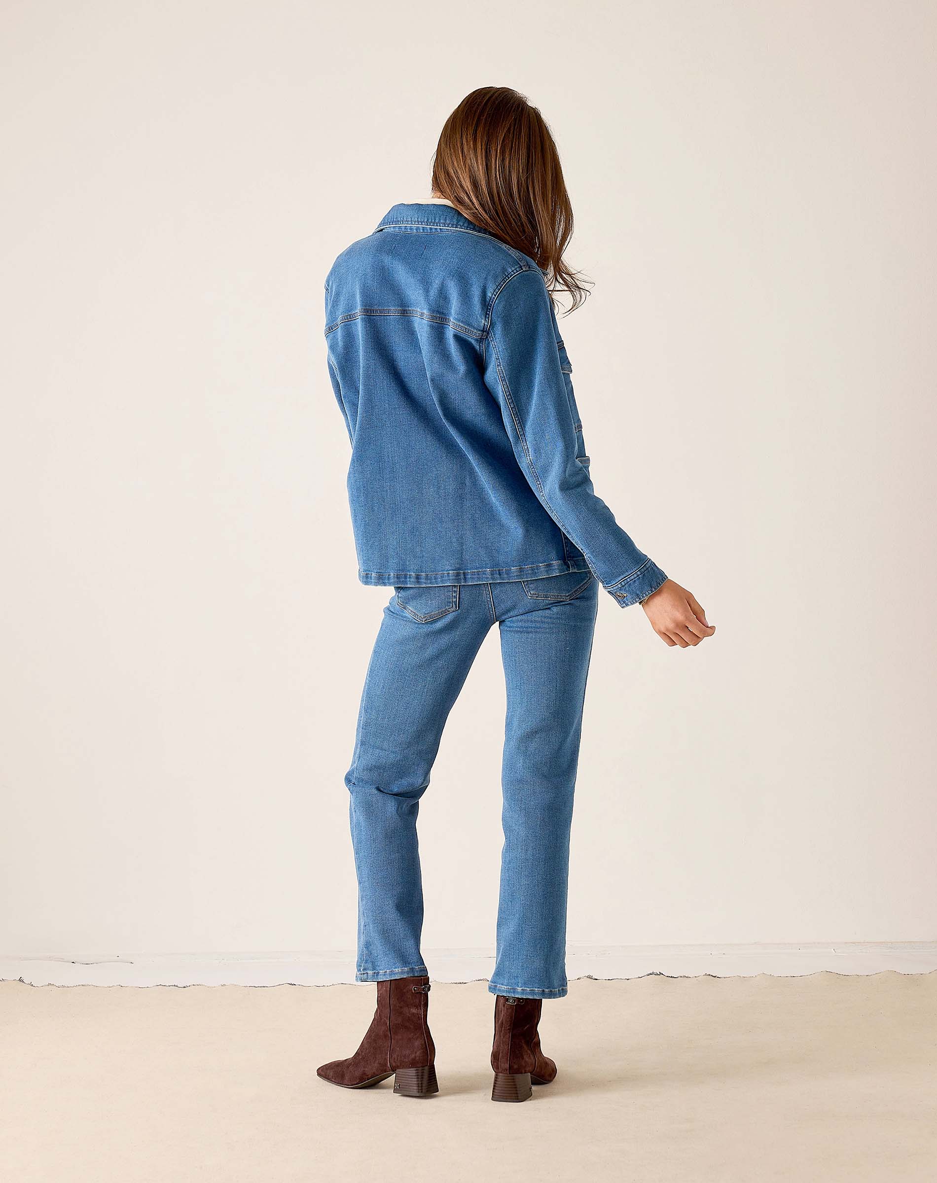 Back view of the medium blue denim jacket and matching jeans with a model in brown boots.