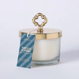 MERSEA ladies' travel-ready fireside Flannel scented soy candle in a clear glass jar with a gold-toned lid featuring an ornate knotted design. Text: 'MERSEA FIRESIDE FLANNEL SCENTED SOY CANDLE MADE IN THE USA'