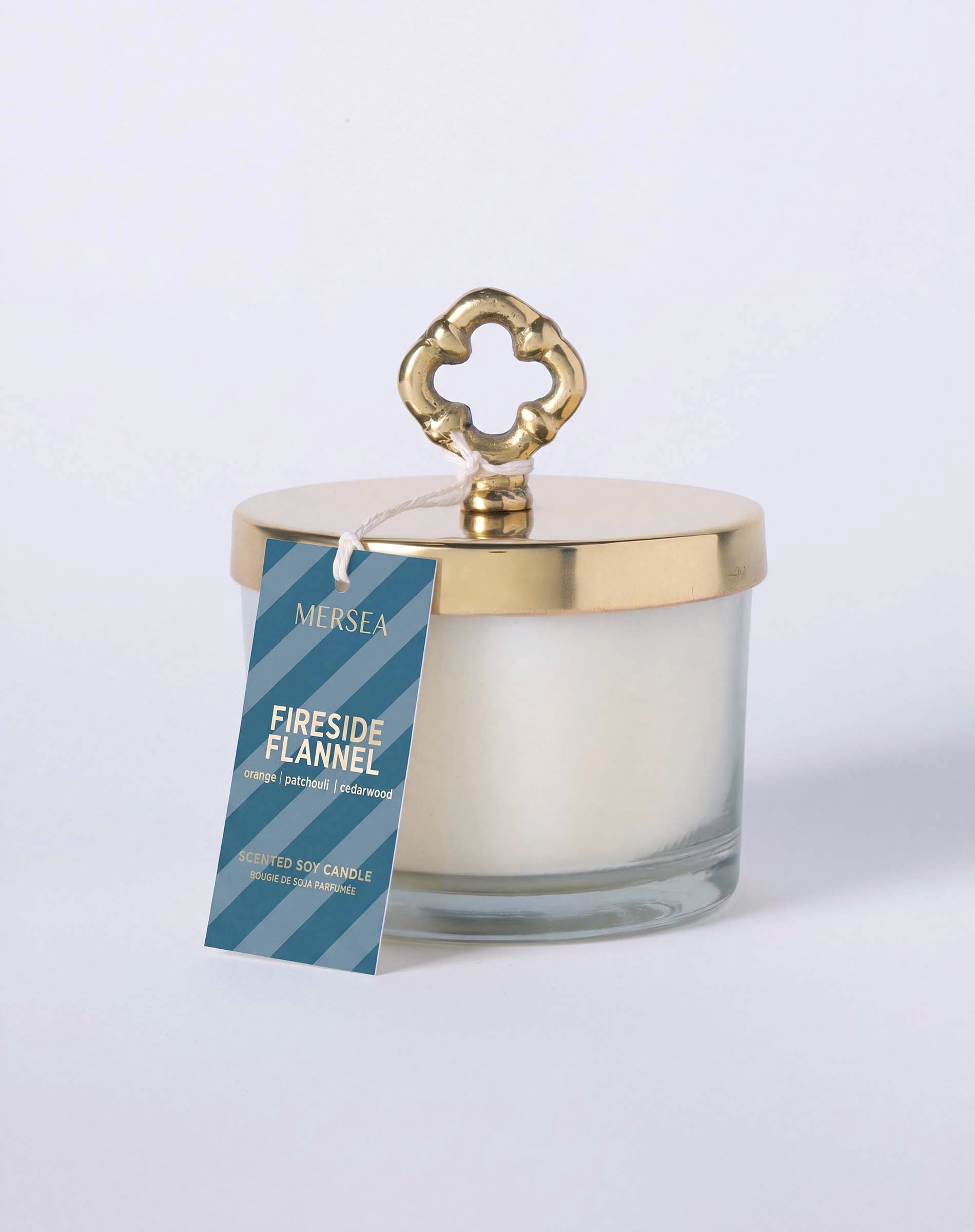 MERSEA ladies' travel-ready fireside Flannel scented soy candle in a clear glass jar with a gold-toned lid featuring an ornate knotted design. Text: 'MERSEA FIRESIDE FLANNEL SCENTED SOY CANDLE MADE IN THE USA'