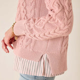 Close-up of a pink cable knit sweater worn over a striped shirt and jeans on a plain background