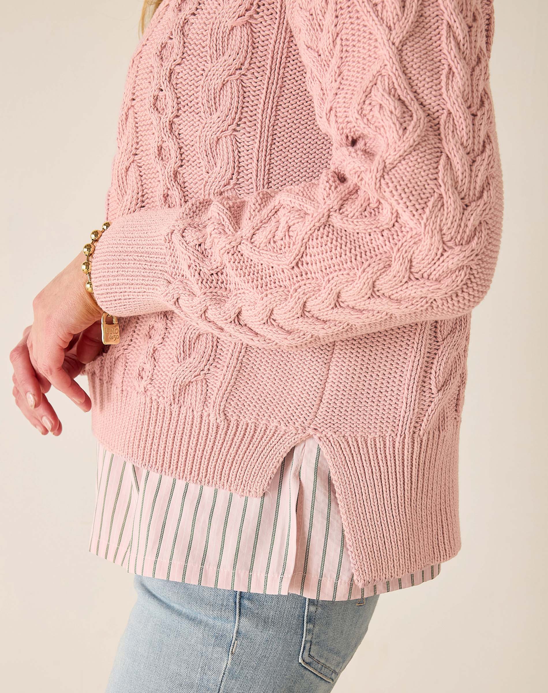 Close-up of a pink cable knit sweater worn over a striped shirt and jeans on a plain background