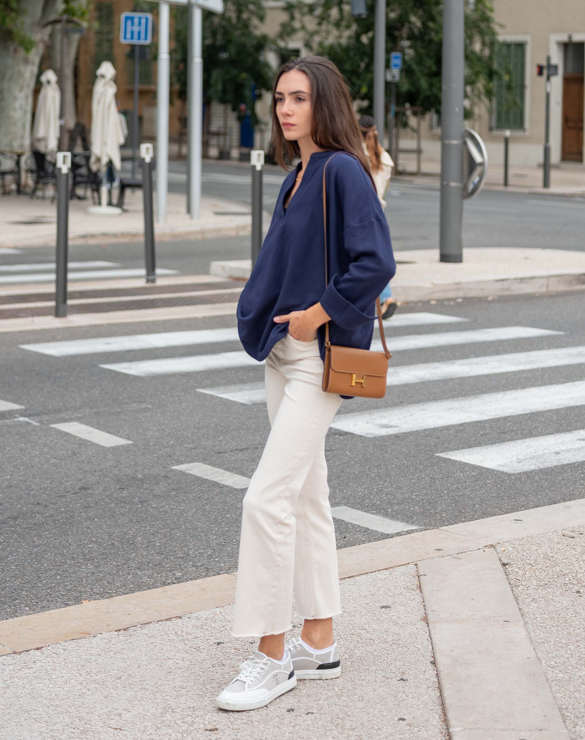 Woman crossing a street wearing a navy sweater, white pants, and white sneakers.