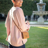 Woman in a pink blouse with a brown handbag standing outdoors with a fountain in the background