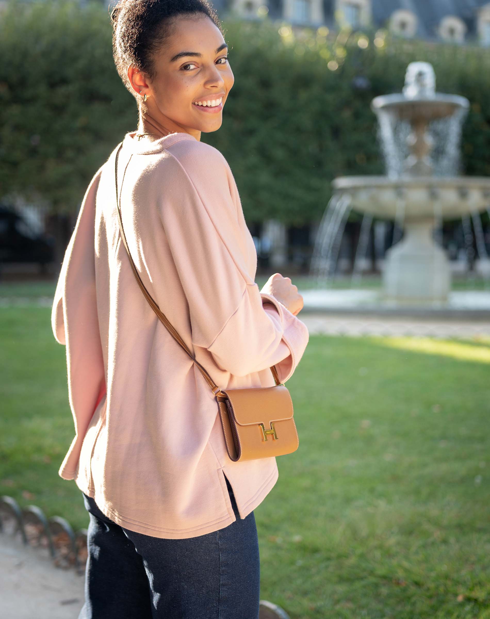 Woman in a pink blouse with a brown handbag standing outdoors with a fountain in the background