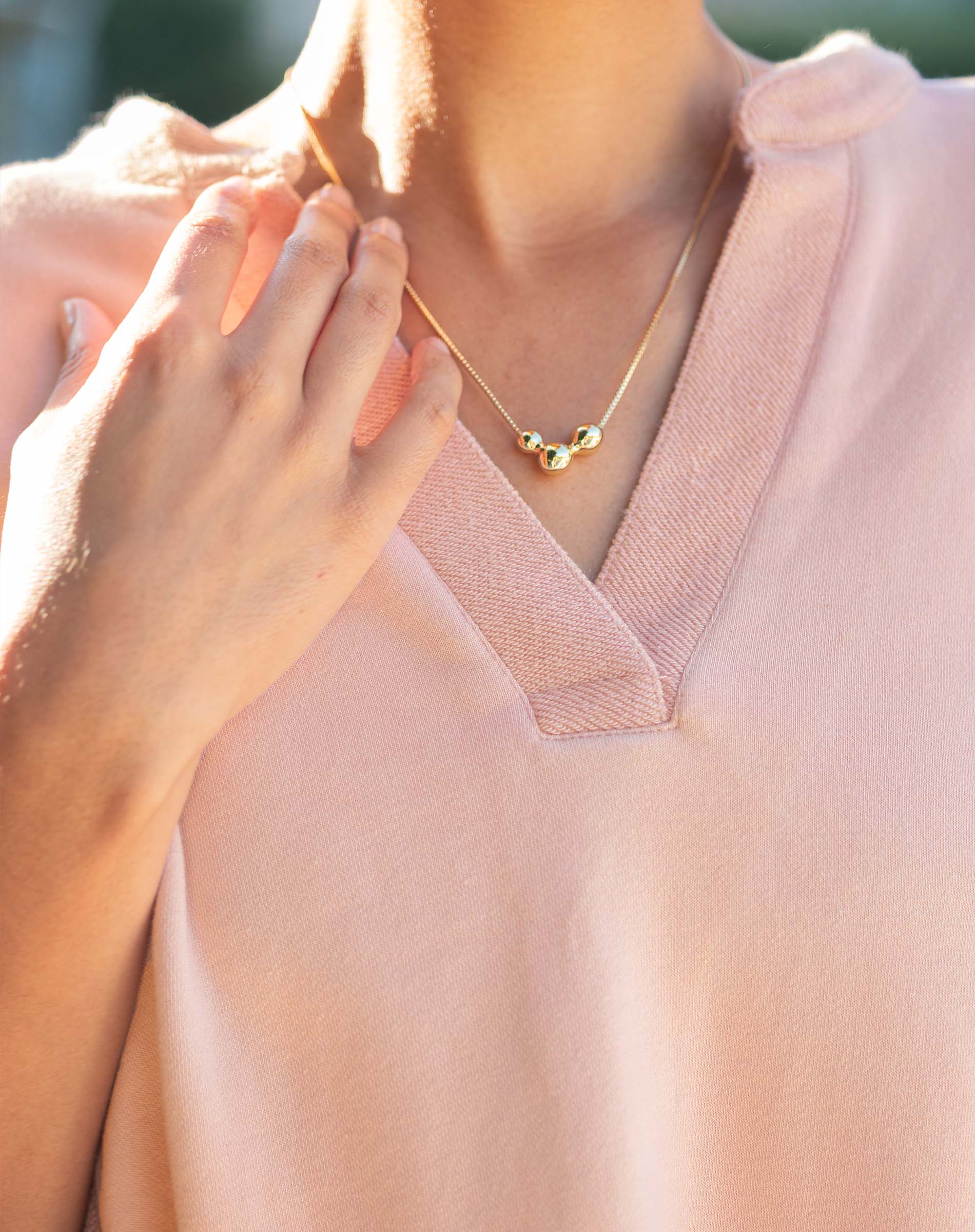 Person wearing a gold necklace with three beads on a light pink top.