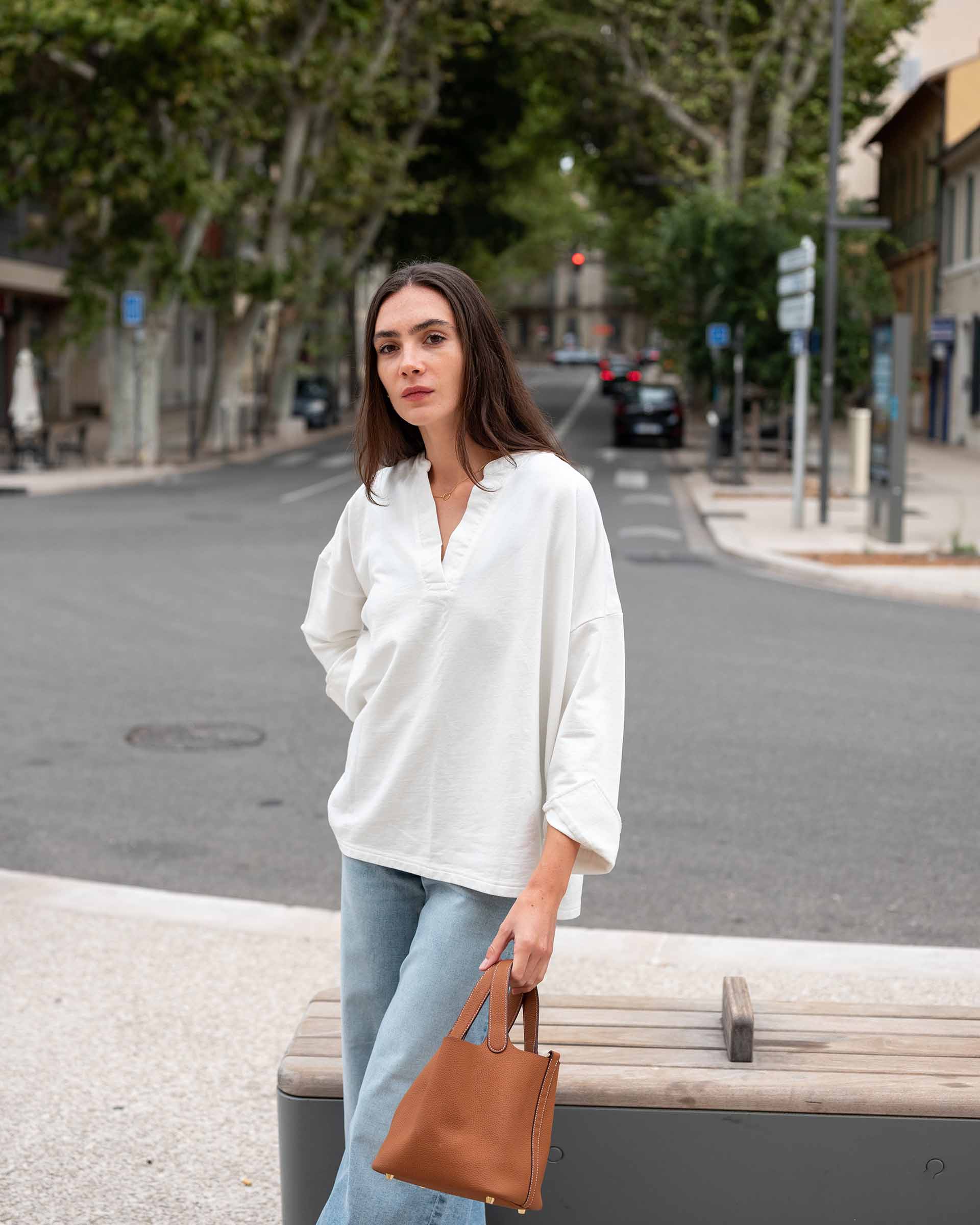 Woman in a white shirt and light blue jeans standing on a street with a brown bag.