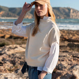 Bone dolman top with a high neck and long sleeves, worn over denim jeans on a rocky beach.