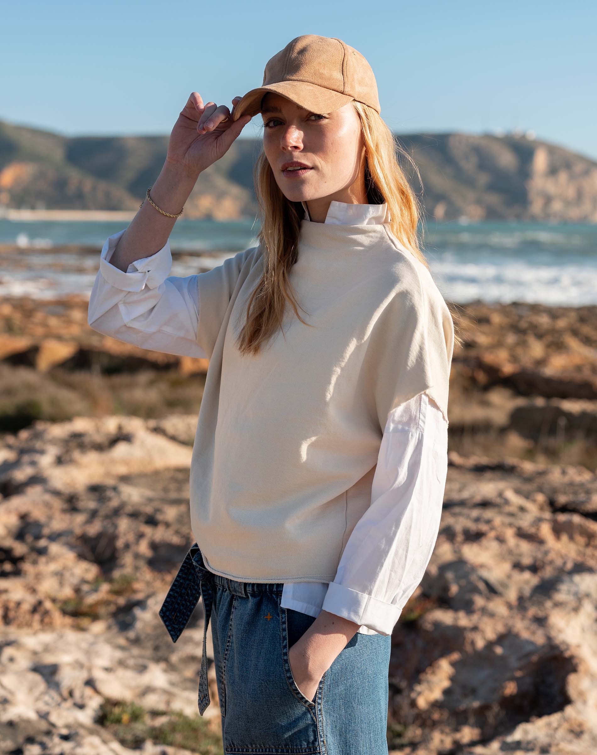Bone dolman top with a high neck and long sleeves, worn over denim jeans on a rocky beach.