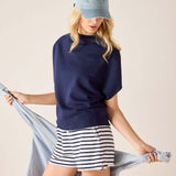 Woman wearing a navy blue top and striped shorts, holding a light gray scarf against a beige background.