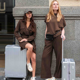 MERSEA women's travel-friendly espresso dolman top and wide-leg trousers worn by two women with suitcases.