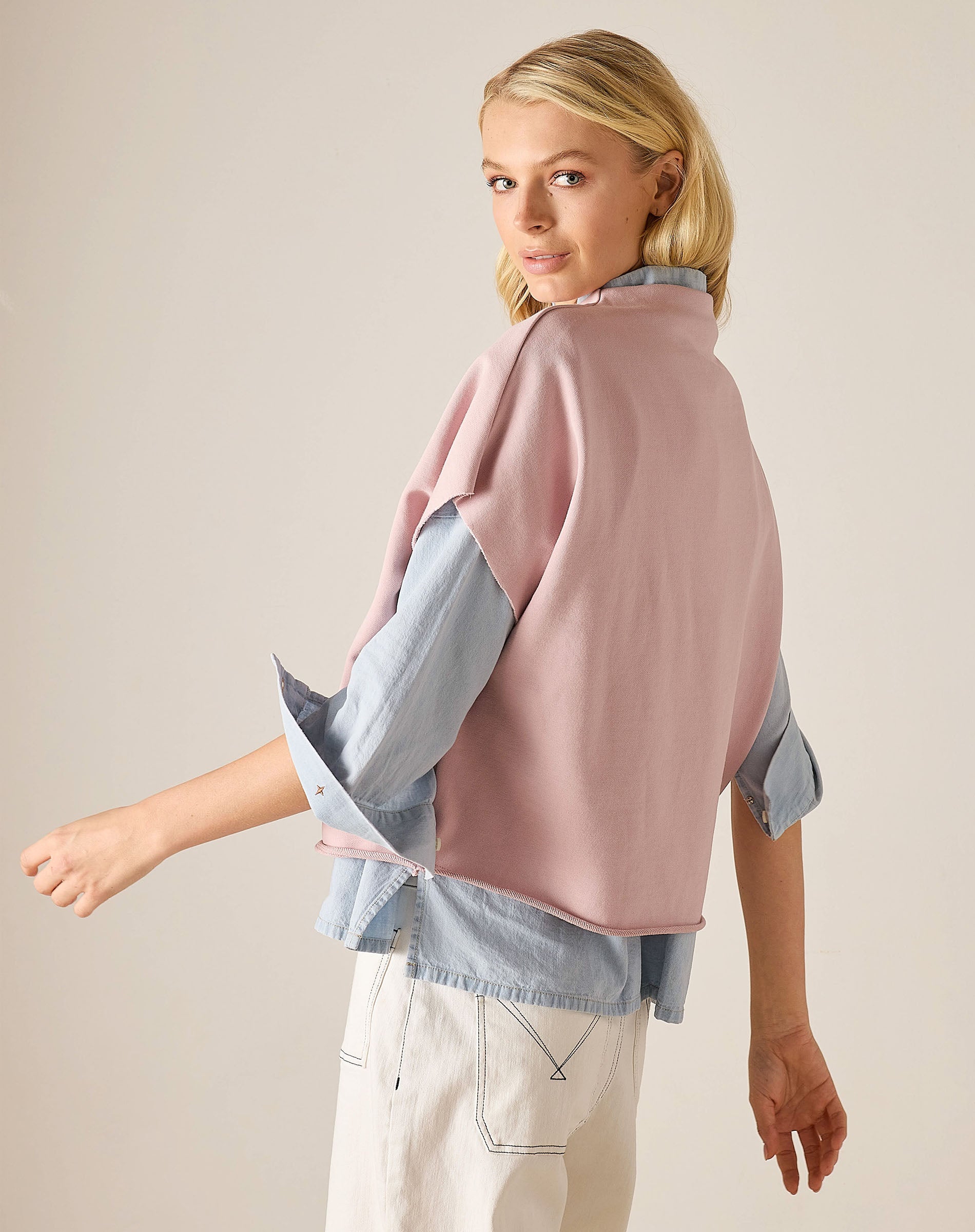 Woman wearing a pink sweater over a blue shirt with beige pants on a plain background