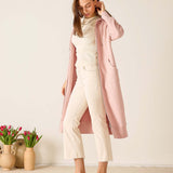 Woman wearing a long pink coat over a white outfit, standing in a room with decorative vases and tulips.