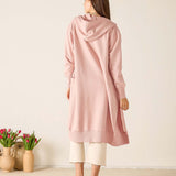 Woman wearing a pink hooded cardigan standing in a minimalistic room with two vases of tulips.
