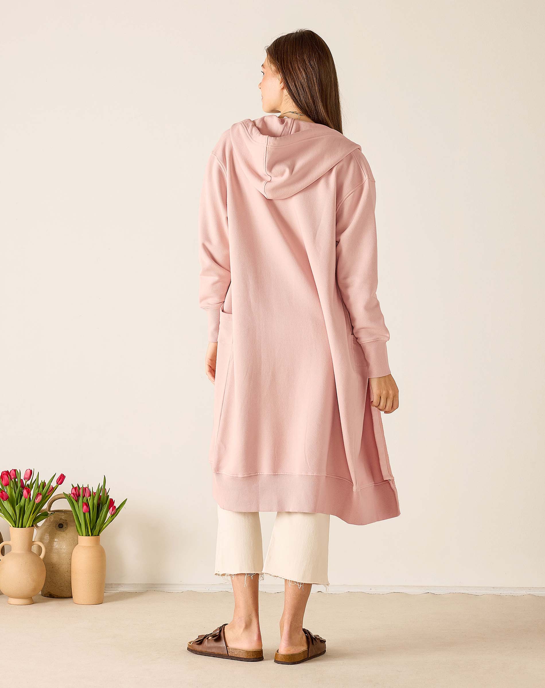 Woman wearing a pink hooded cardigan standing in a minimalistic room with two vases of tulips.