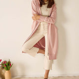Woman wearing a pink cardigan over a white outfit with a vase of flowers on the floor.
