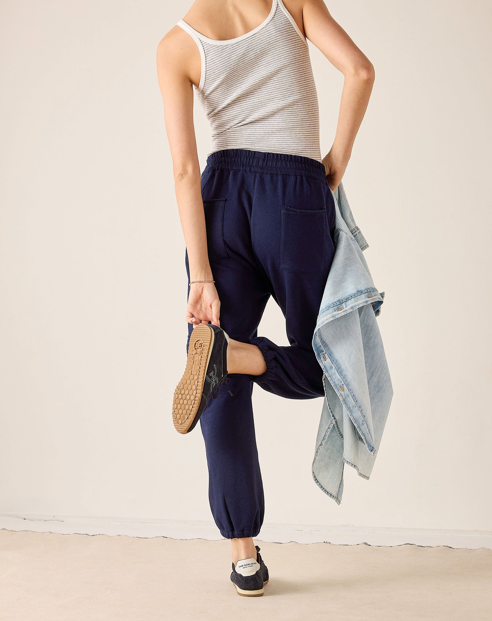 Person wearing navy jogger pants and a striped tank top, holding a denim jacket.