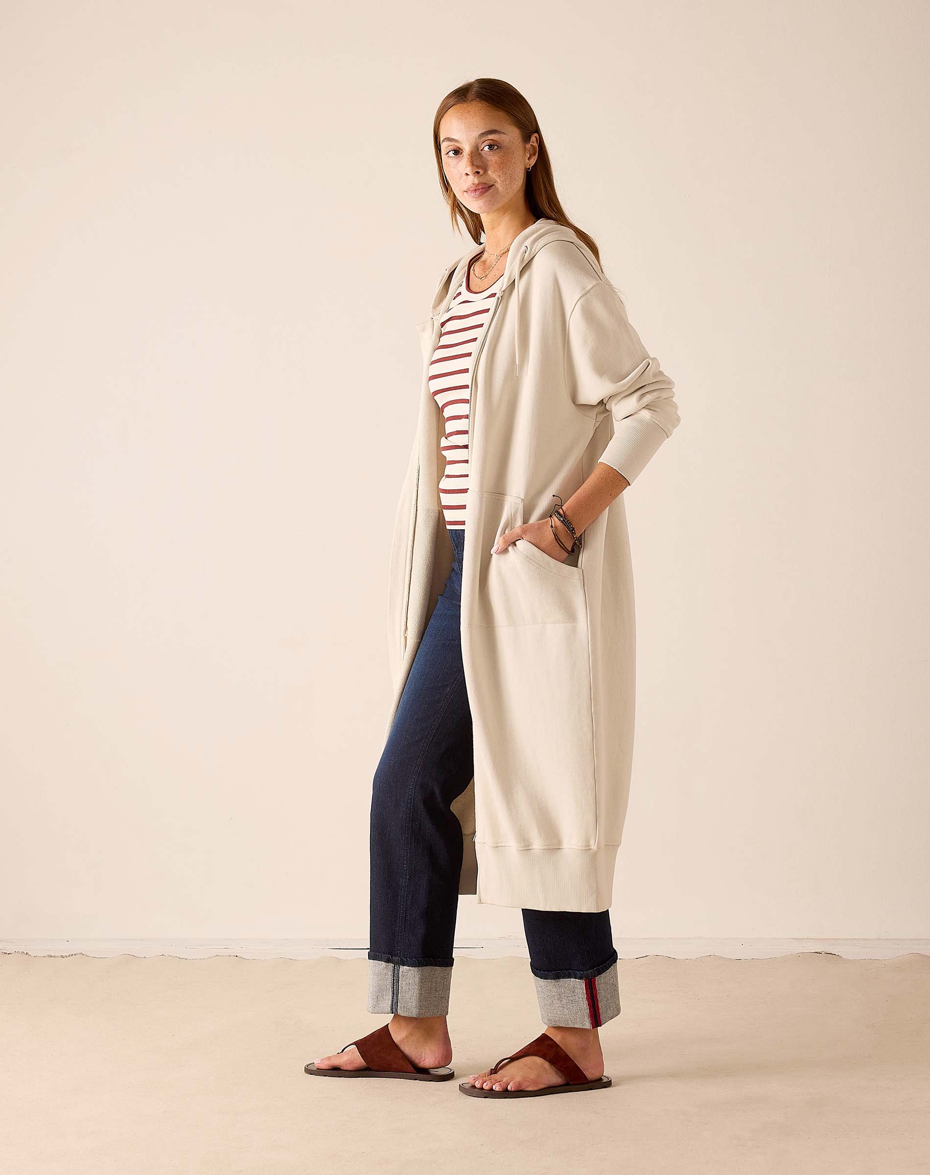 Bone hoodie with a hood and front pocket over a striped tee and dark trousers.