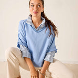 Woman wearing a light blue sweater over a striped shirt with beige pants on a plain background