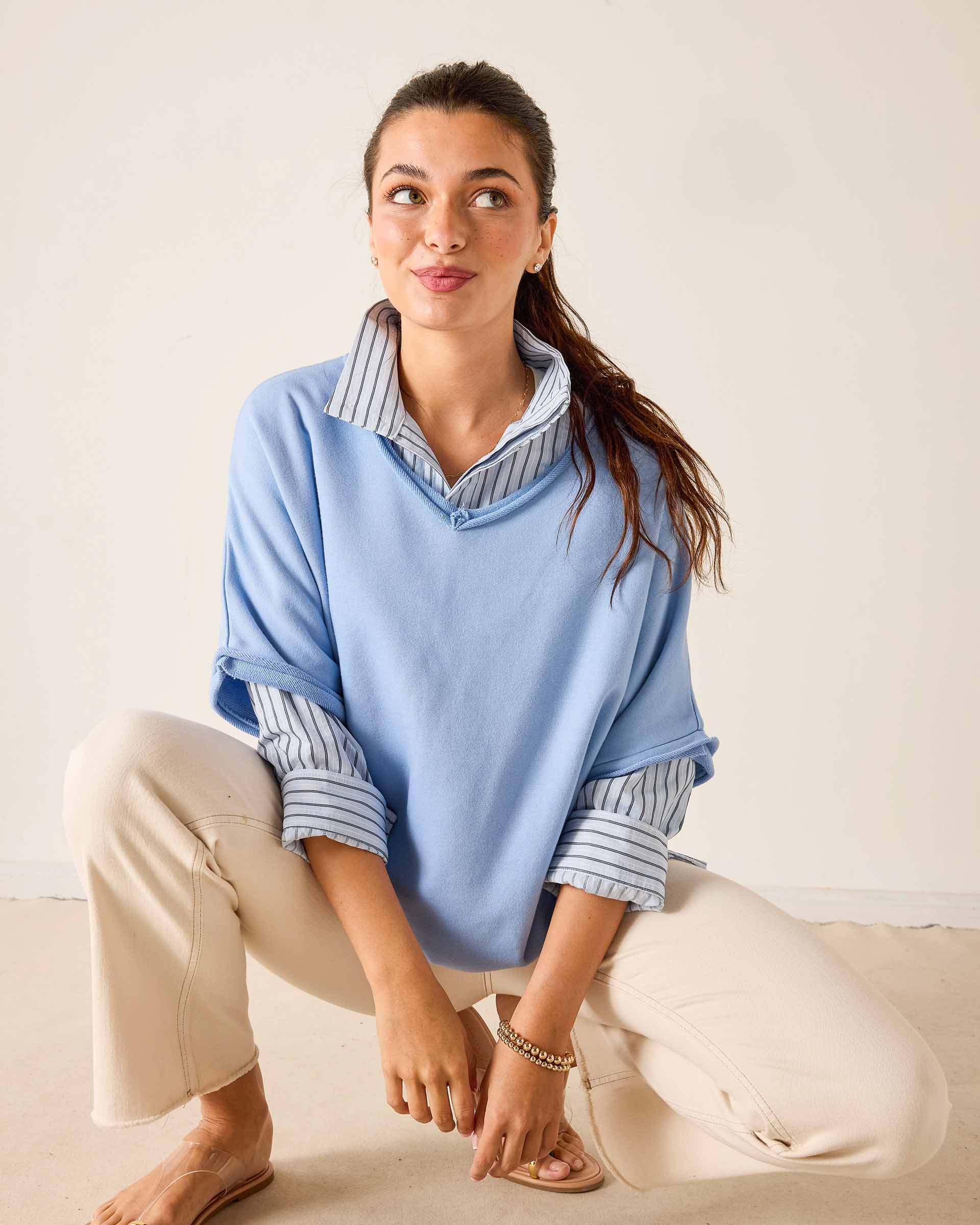 Woman wearing a light blue sweater over a striped shirt with beige pants on a plain background