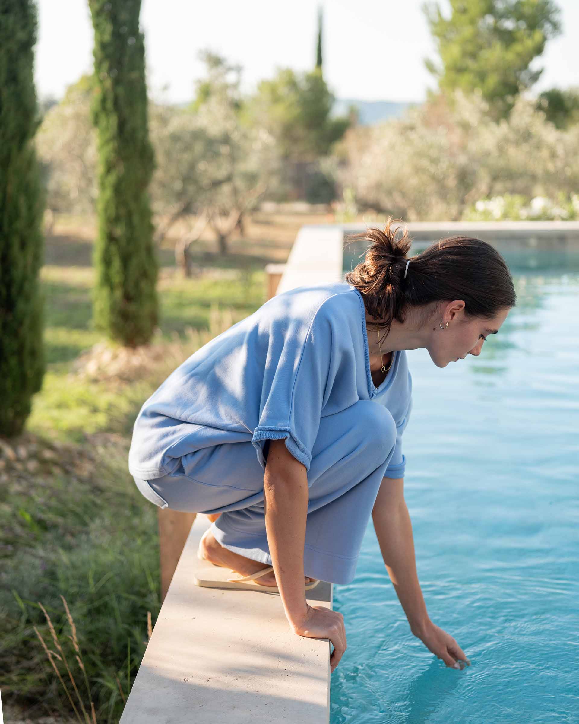Woman in a blue robe standing by a pool with greenery in the background