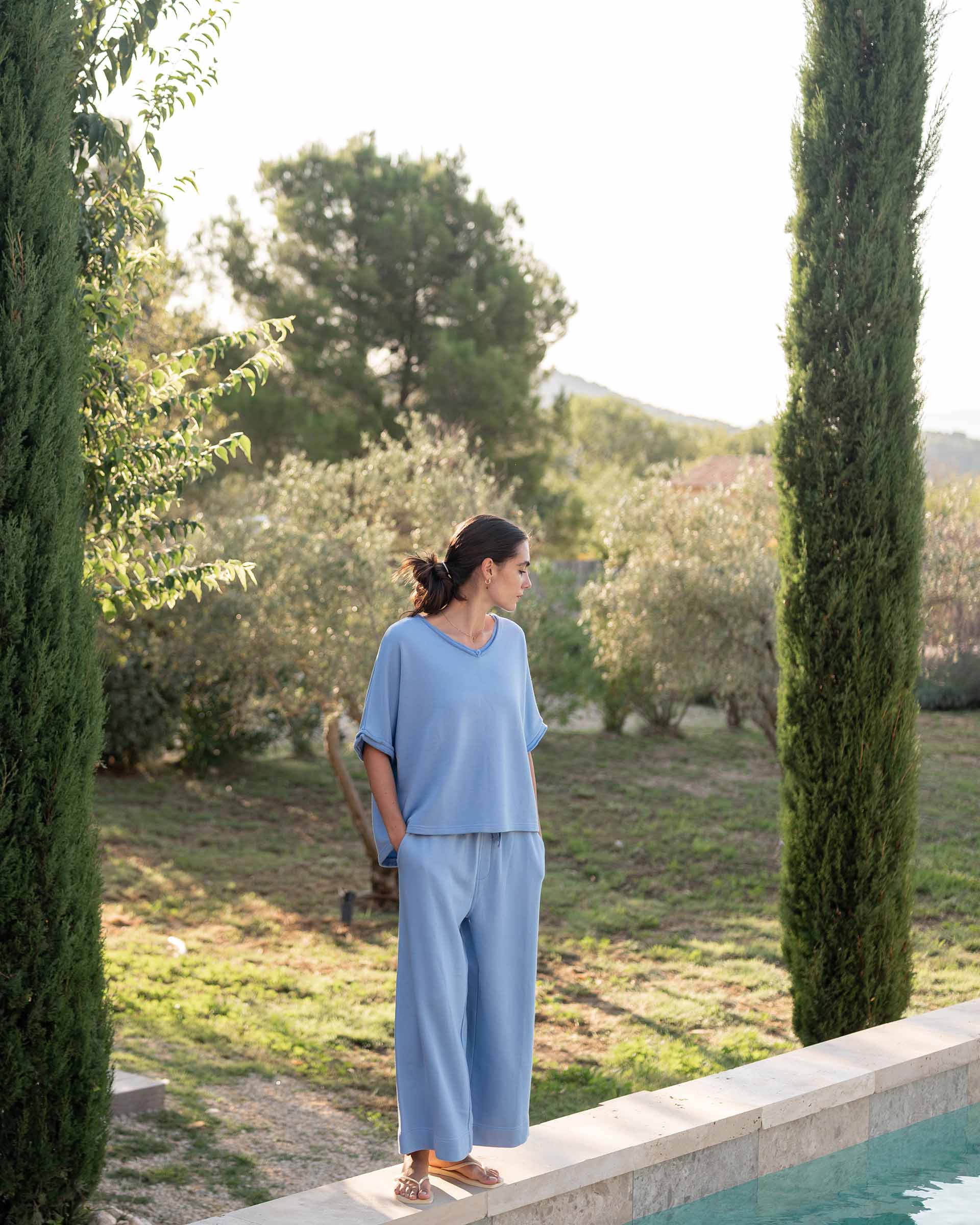 Woman in a light blue outfit standing by a pool with greenery in the background