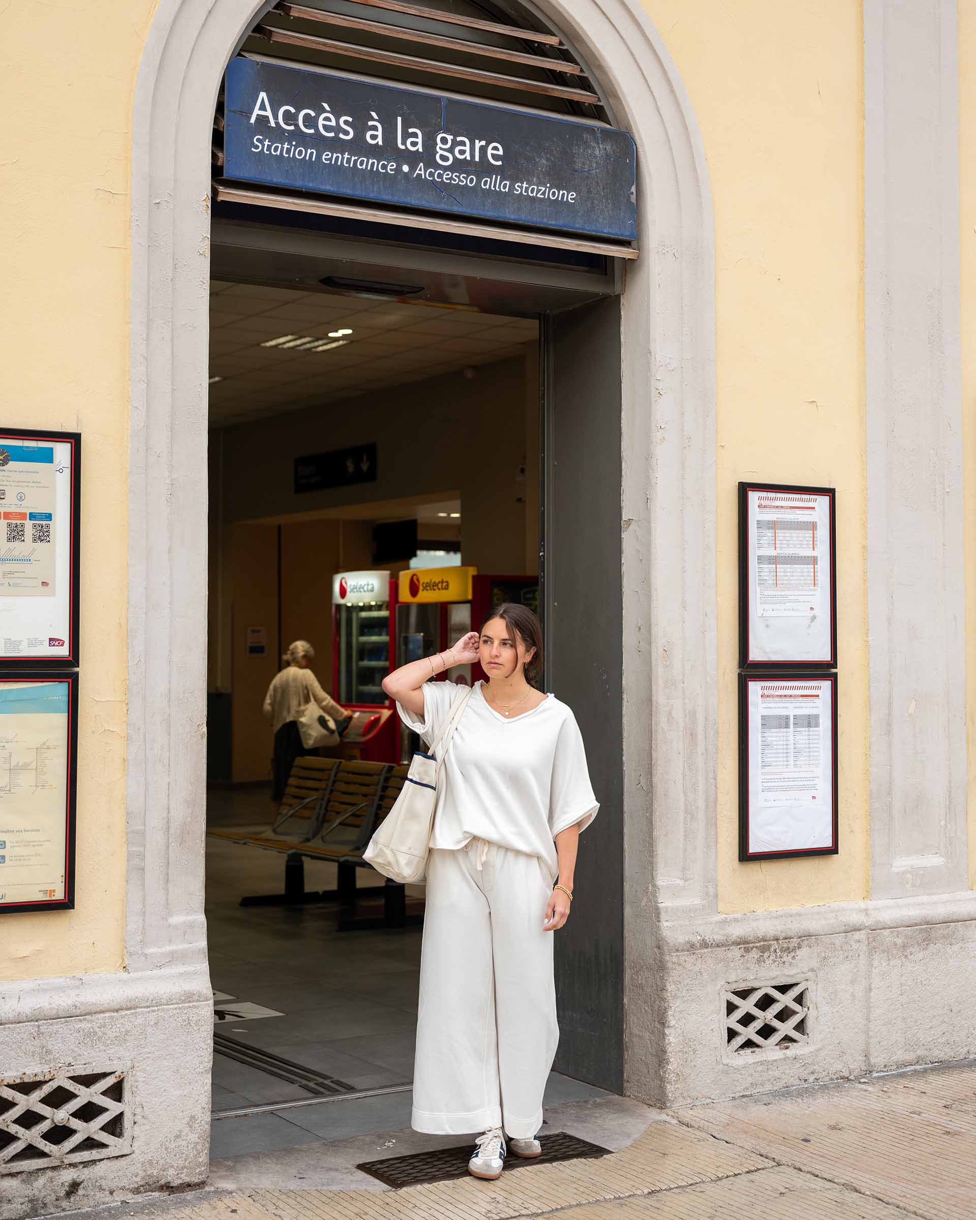 Woman in a white outfit standing in front of a station entrance with 'Accès à la gare' sign.