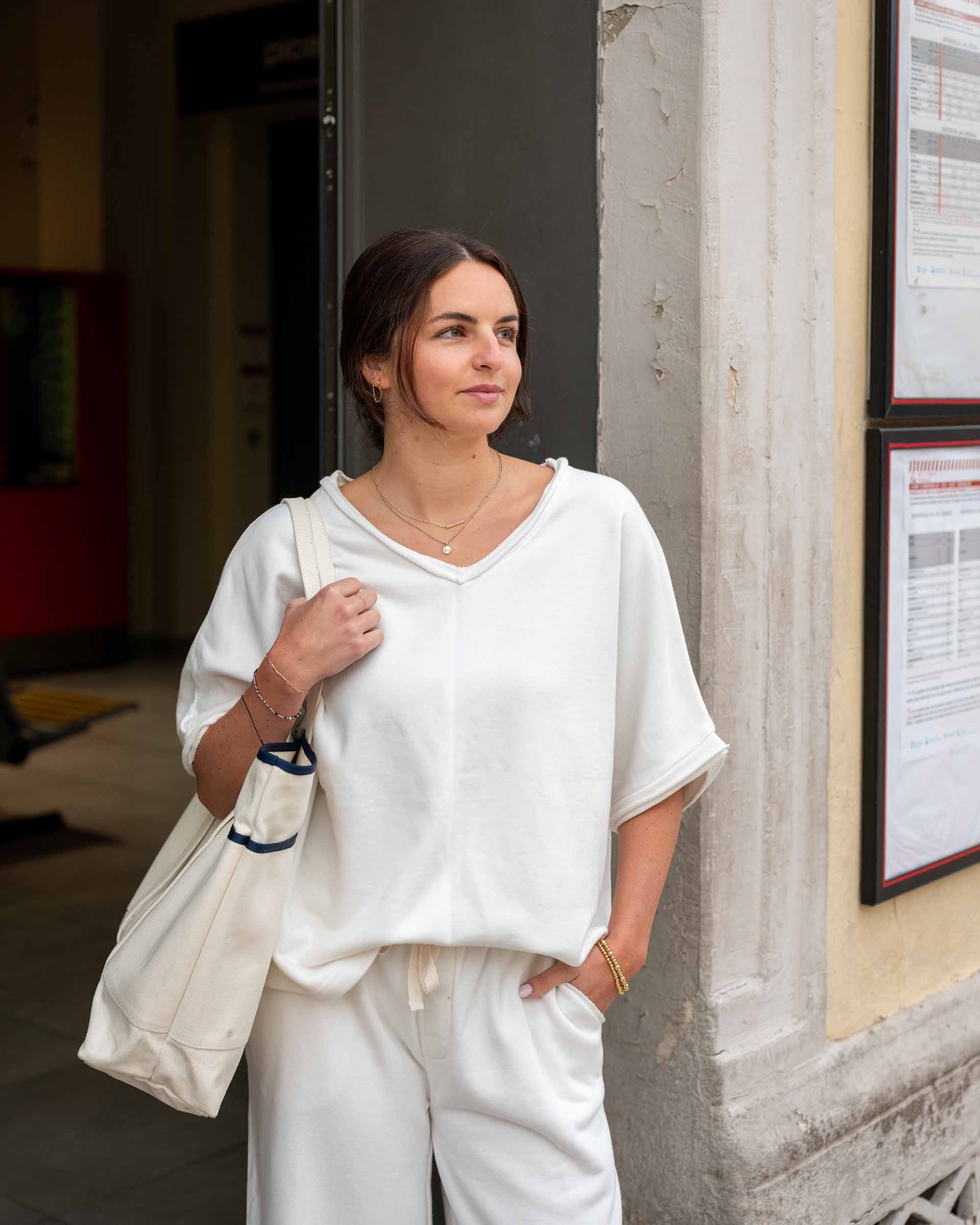Woman in white outfit holding a tote bag, standing against a building wall.