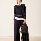 Person wearing a black sweater and pants with a white shirt, holding a black backpack on a beige floor.