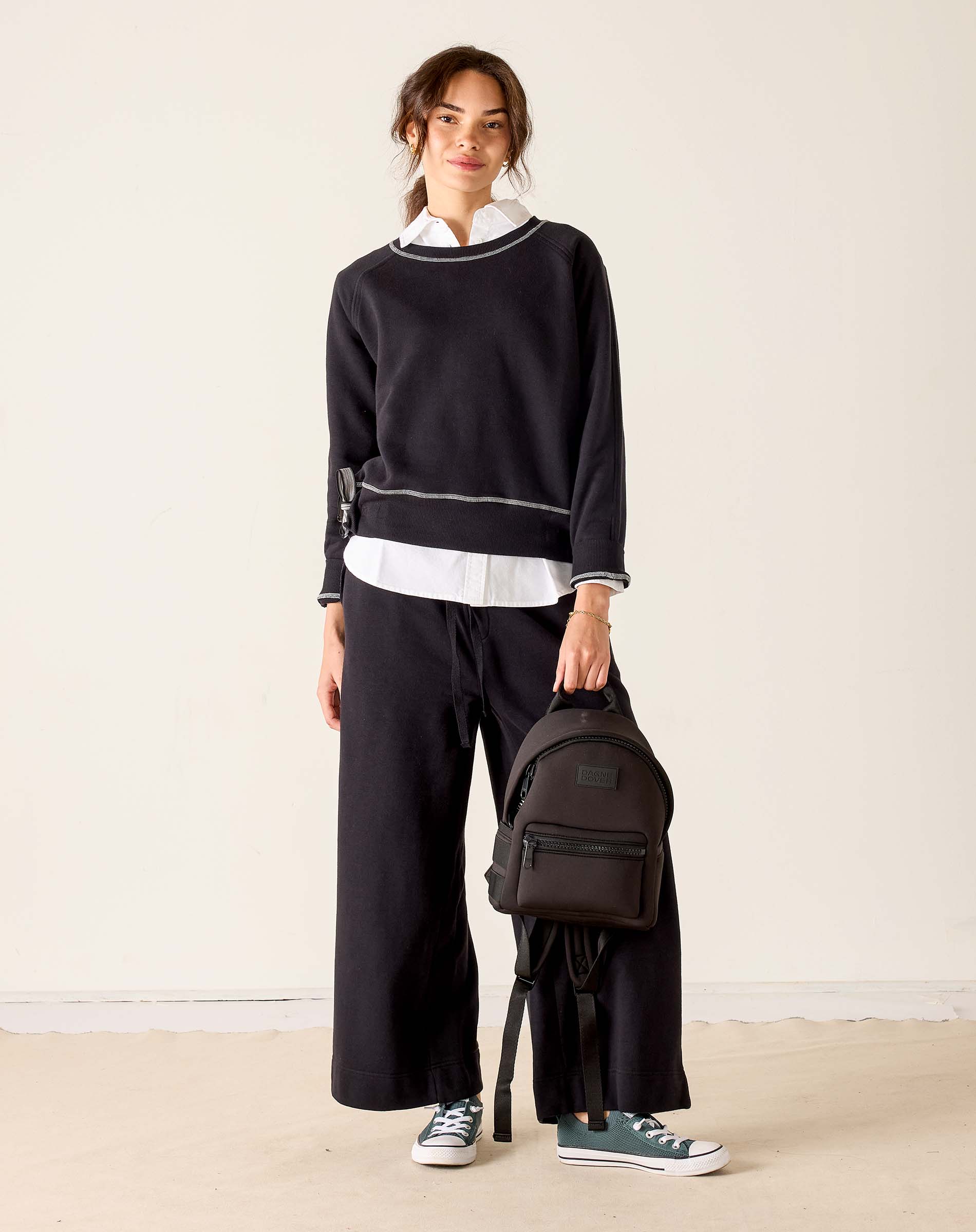 Person wearing a black sweater and pants with a white shirt, holding a black backpack on a beige floor.