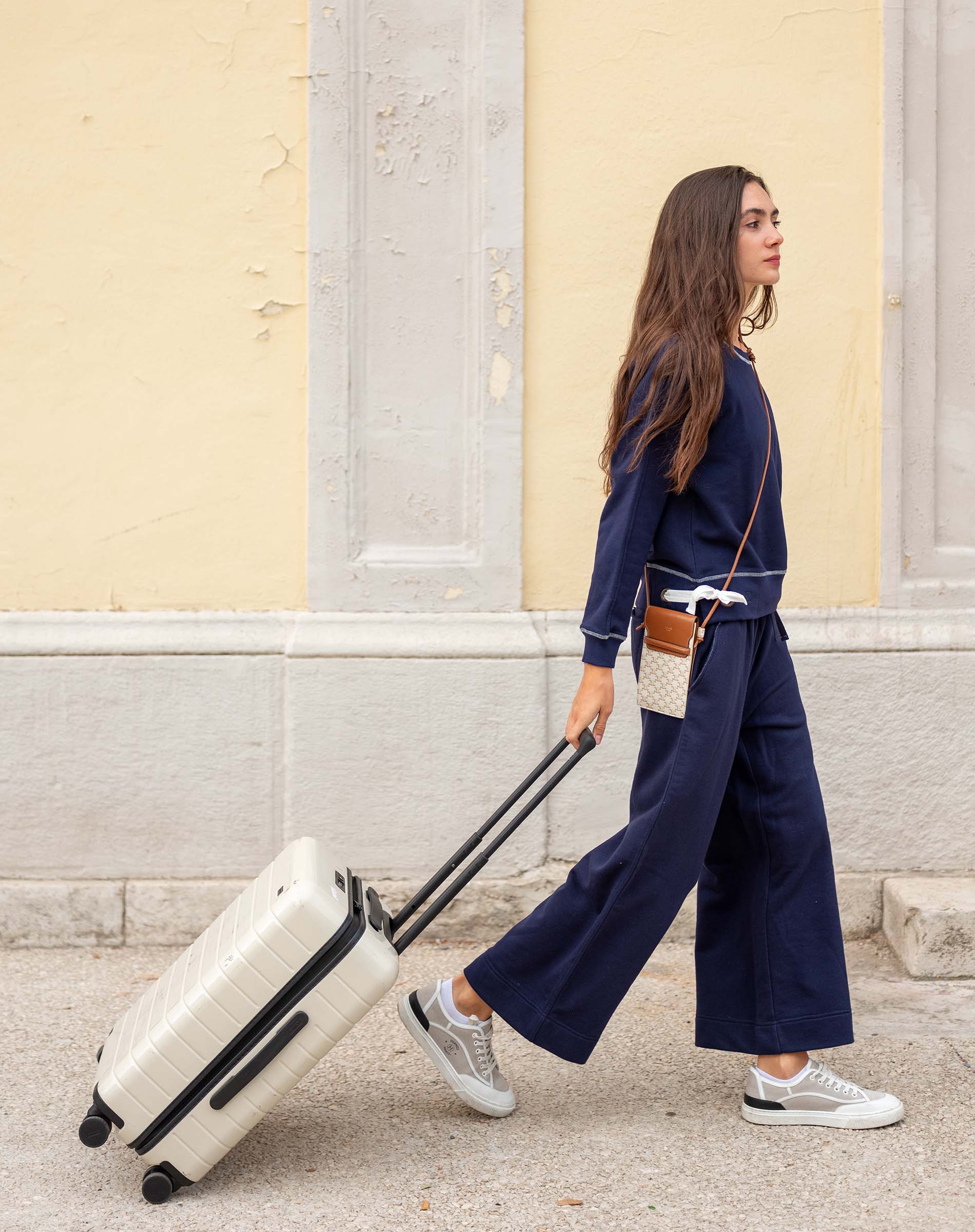 Woman in navy outfit pulling a white suitcase on a street.