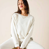 Woman wearing a white sweatshirt and pants sitting on a wooden stool against a beige wall.