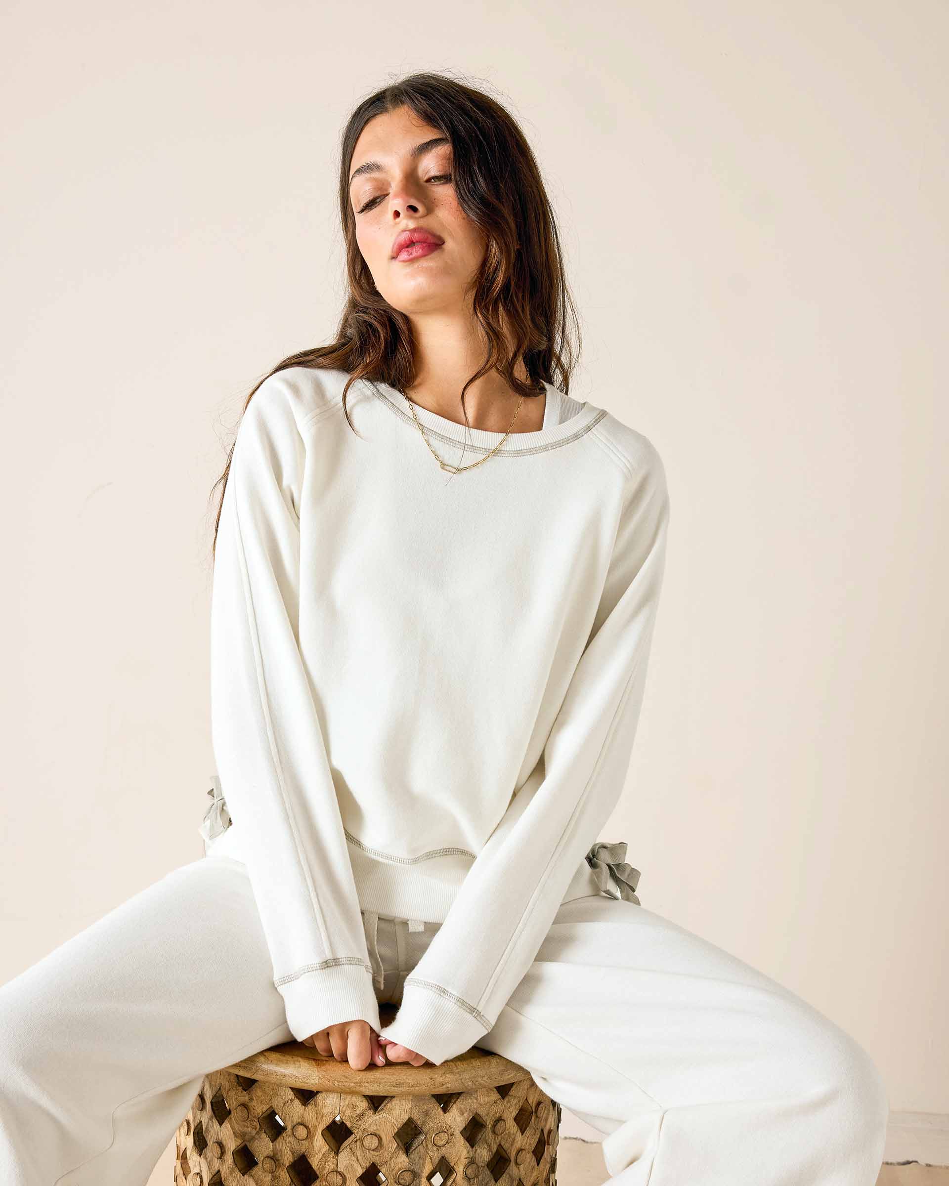 Woman wearing a white sweatshirt and pants sitting on a wooden stool against a beige wall.